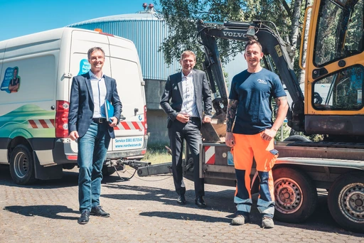  Drei Männer stehen im Freien vor einem weißen Transporter und einem gelben Bagger