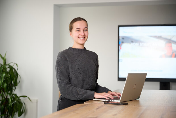 Eine junge Frau steht am Tisch, vor ihr ein aufgeklappter Laptop, hinter ihr ein Monitor an der Wand.